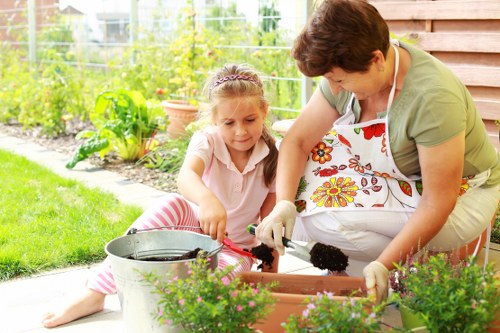 A woman and a young girl are working together in a well-maintained garden, engaging in gardening activities near a wooden shed. The woman, wearing an apron and gardening gloves, is helping the girl plant or tend to a flower bed that contains vibrant, flowering plants with green foliage. The girl, dressed in a light pink top and striped trousers, is holding a gardening tool and appears focused on the task. In front of them, there is a metallic bucket, possibly for watering or collecting garden waste. The garden features a lush, green lawn in the foreground, and behind them is a raised flower bed bordered by soil and some small green plants. The background shows a wire fence and a variety of leafy bushes and plants, indicating an outdoor space in a suburban or residential area, possibly within Manor Park. The scene is captured on a bright, sunny day, with natural light illuminating the garden, highlighting the natural tones of the plants, soil, and wooden structures, reflecting typical garden maintenance work by professional gardening services such as Hedge Trimming Manor Park.