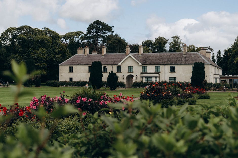 A historic brick mansion with multiple chimneys and a slate roof, surrounded by well-maintained garden beds and lush greenery. The garden features neatly trimmed bushes, vibrant flowering plants, and 